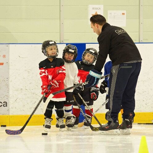 Small children learning how to play hockey, dressed in hockey equipment on the ice with a coach